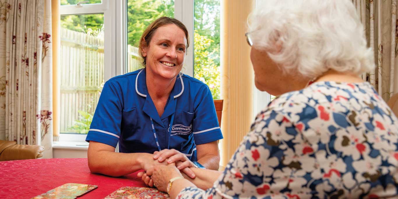 Dementia Care in Basingstoke & Deane female care assistant with female customer in home at kitchen table holding hands