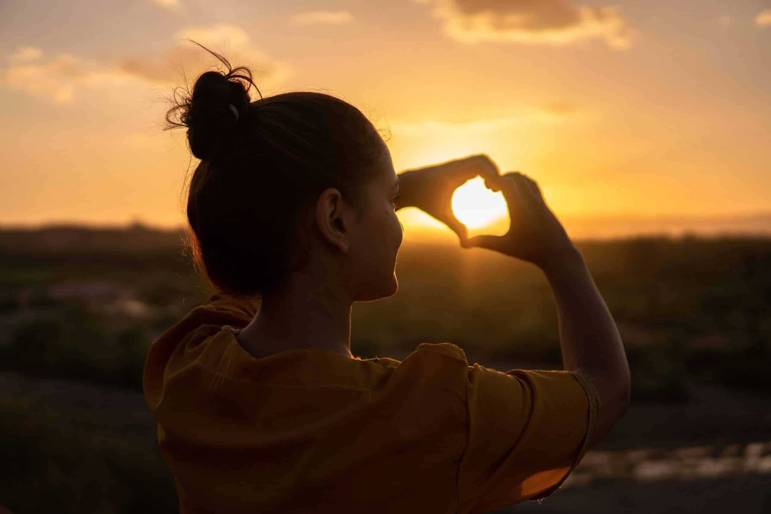 a woman wearing an orange shirt making a love heart with her hands in the sunset for how to manage stress in a healthy way as a carer
