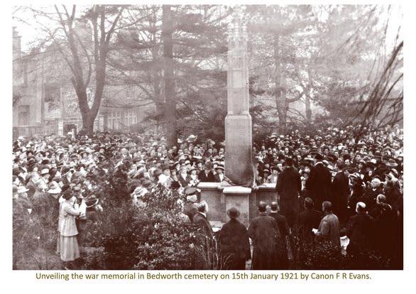 A large crowd gathers around a newly unveiled war memorial cross in Bedworth cemetery on 15th January 1921, captured in a sepia-toned historical photograph. People in early 20th-century attire, including hats and coats, stand solemnly among leafless trees and gravestones, with a woman in the foreground possibly reading or speaking.