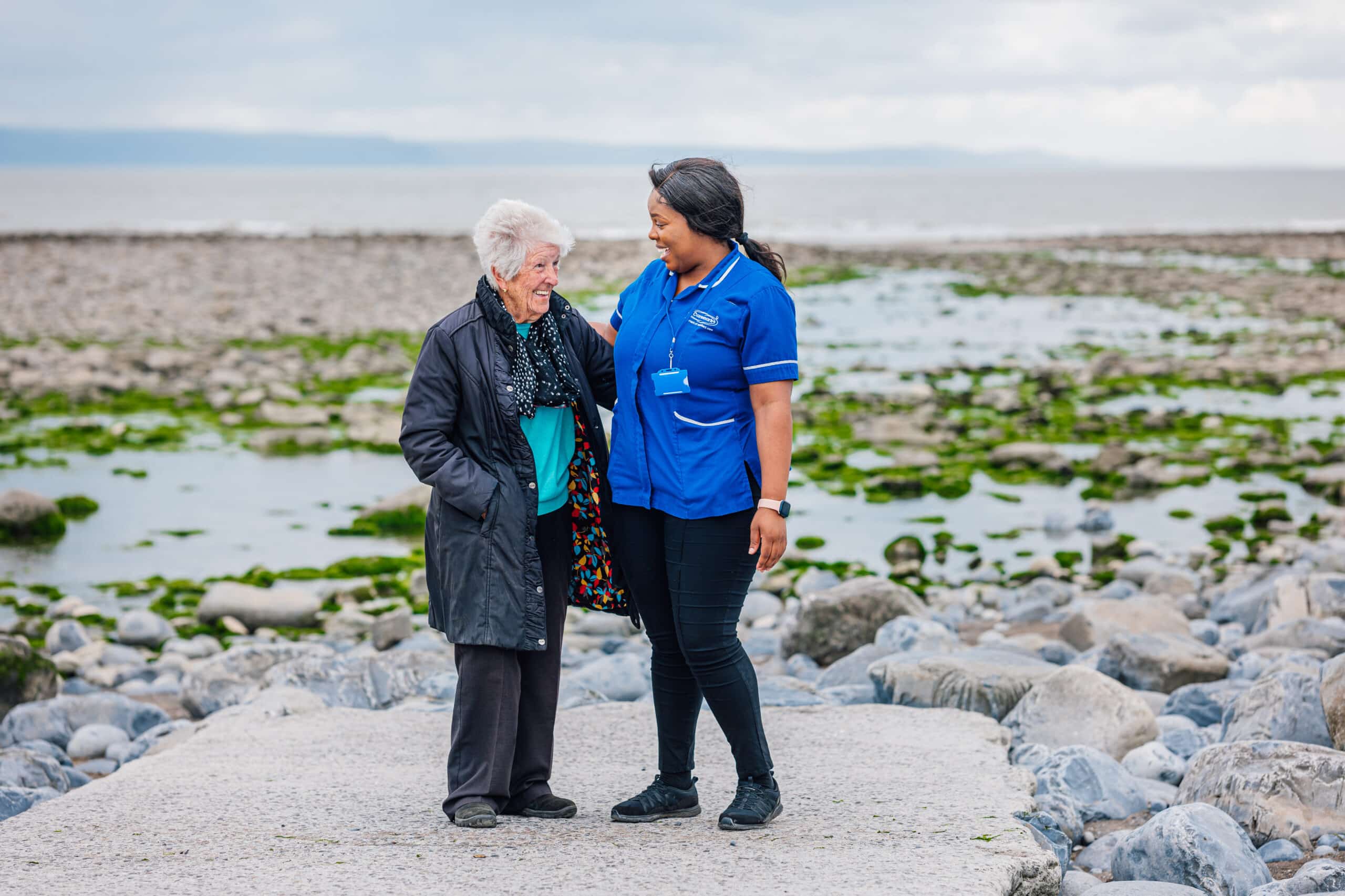 A care worker and an older client standing on the beach, smiling at each other, enjoying a sunny day