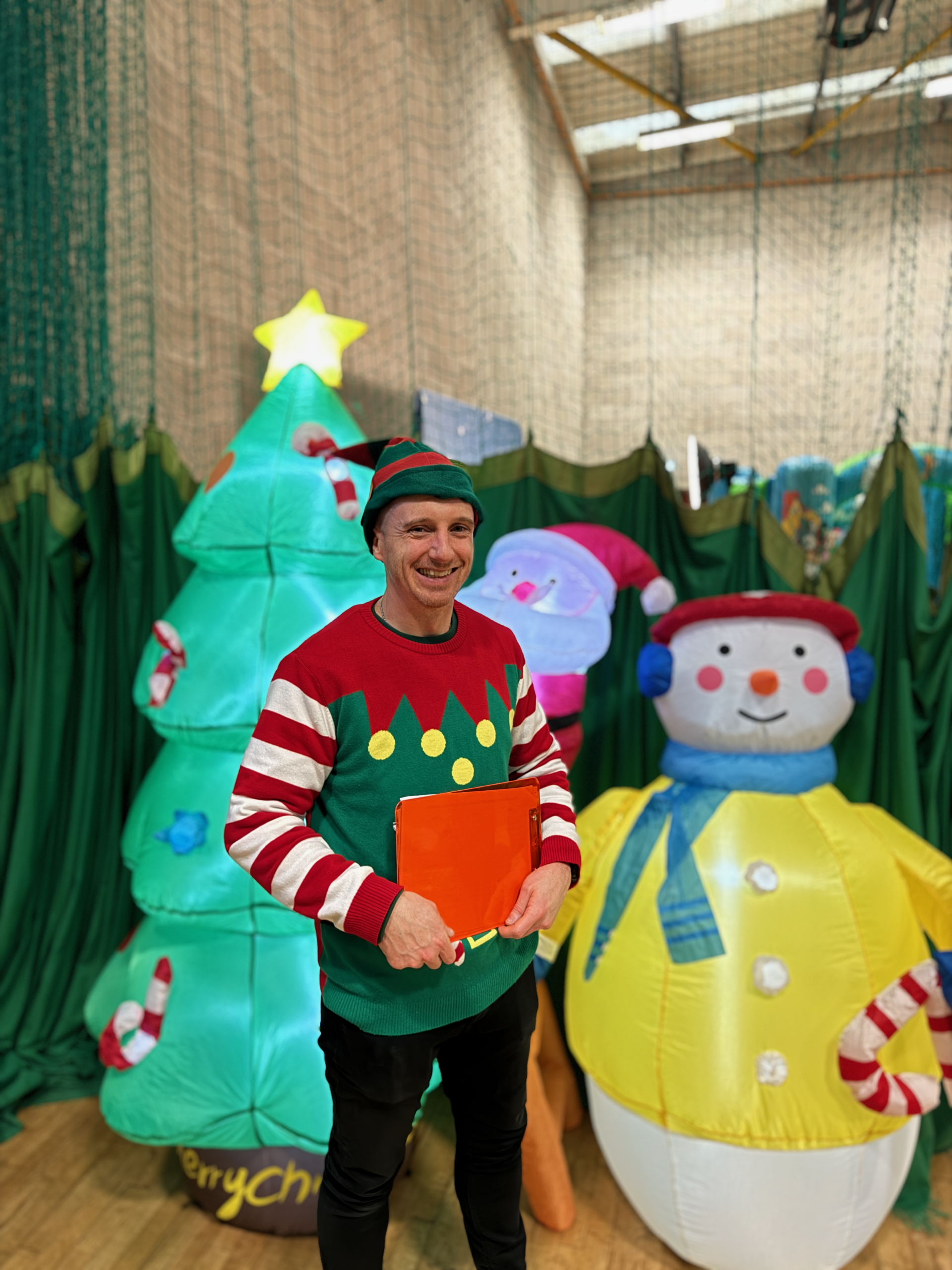 Man dressed as a festive elf, wearing a green and red costume with a hat, smiling at the camera