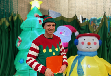 Man dressed as a festive elf, wearing a green and red costume with a hat, smiling at the camera