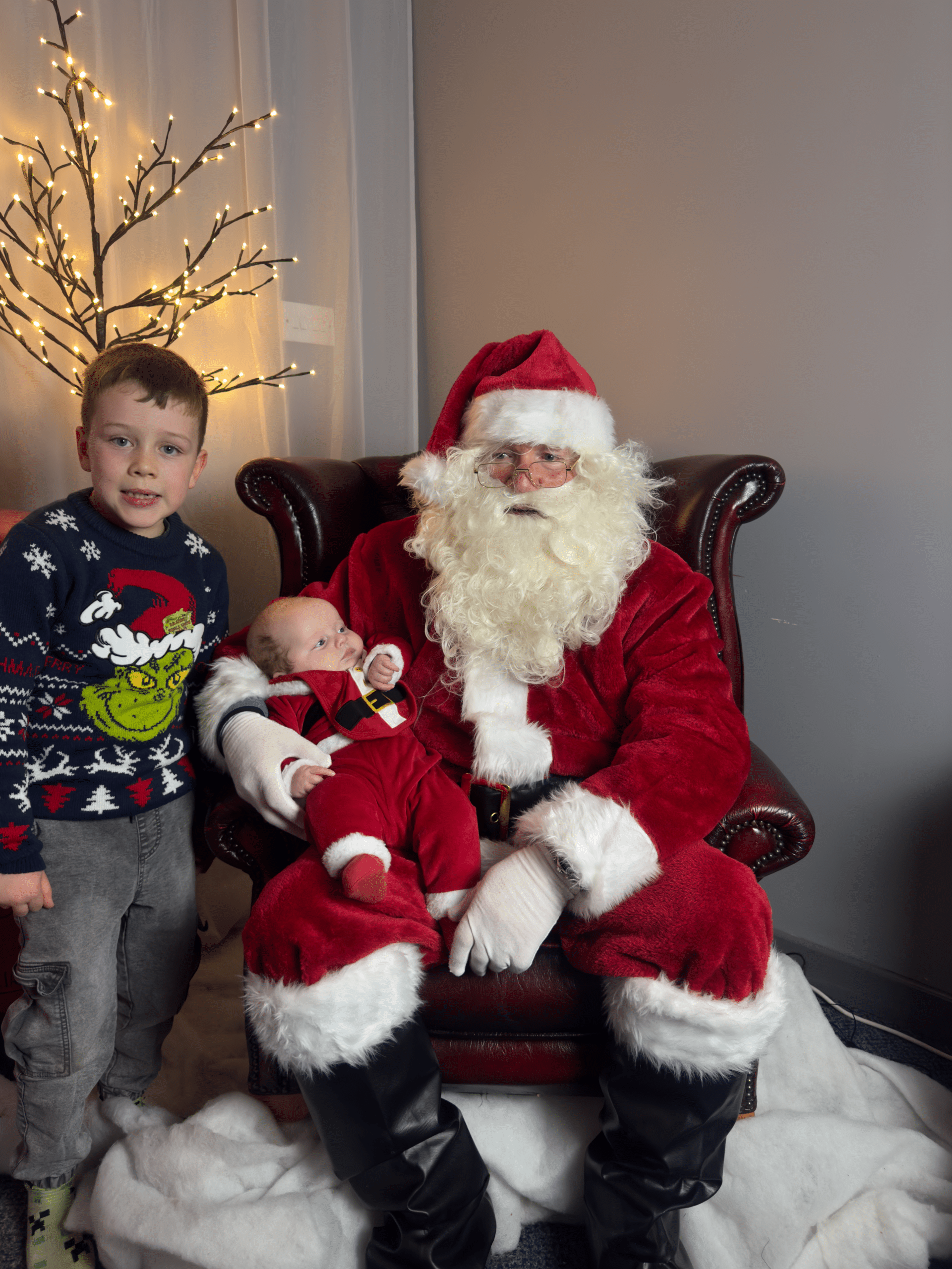 Santa holding a baby dressed in a Santa outfit, while a smiling young boy stands beside them