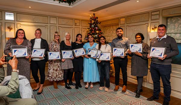 Caremark team awards photo at a Christmas event, with staff holding Caremark certificates in front of a decorated Christmas tree in a hotel function room.