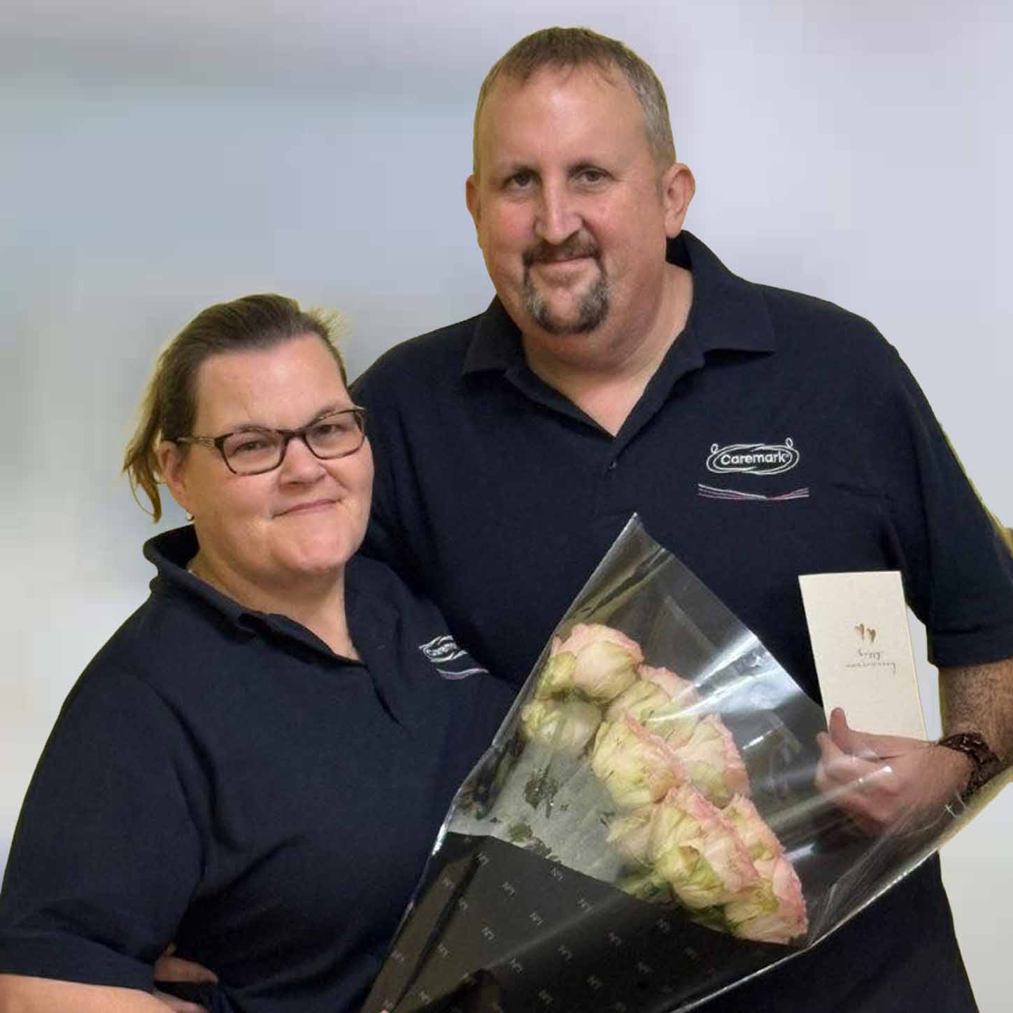 Caremark franchise owners Jo and Paul from Caremark North Dorset and South Wiltshire holding flowers and a card, wearing Caremark polo shirts.
