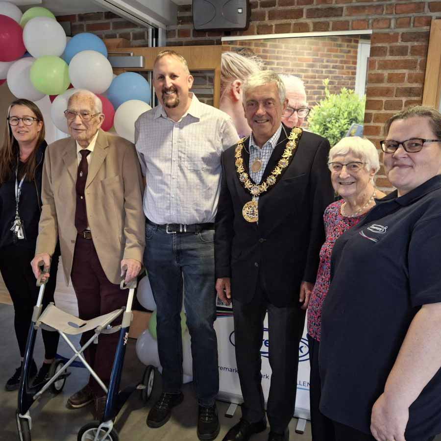 Caremark North Dorset and South Wiltshire community event photo with Caremark team, an elderly couple, and a local mayor, with colourful balloons in the background.