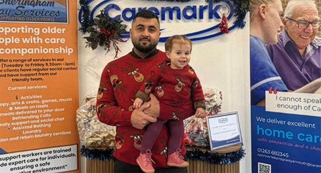 Kawan, franchise owner of Caremark North Norfolk and Great Yarmouth talks about being a young business owner in franchising and why he chose Caremark. Caremark office display with a young business owner holding a child, standing in front of Caremark signage and local home care materials.