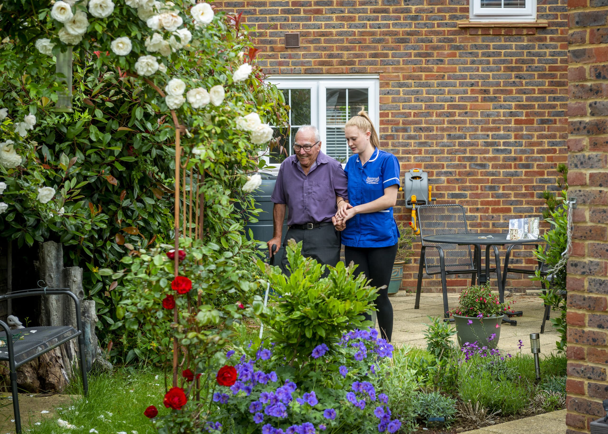 A female care assistant walking arm and arm in a garden with an elderly male service user