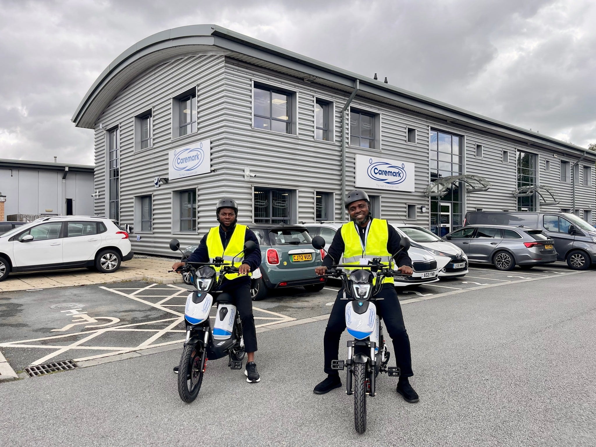 two care assistants on Caremark e-bikes, wearing helmets and high-visability jackets.
