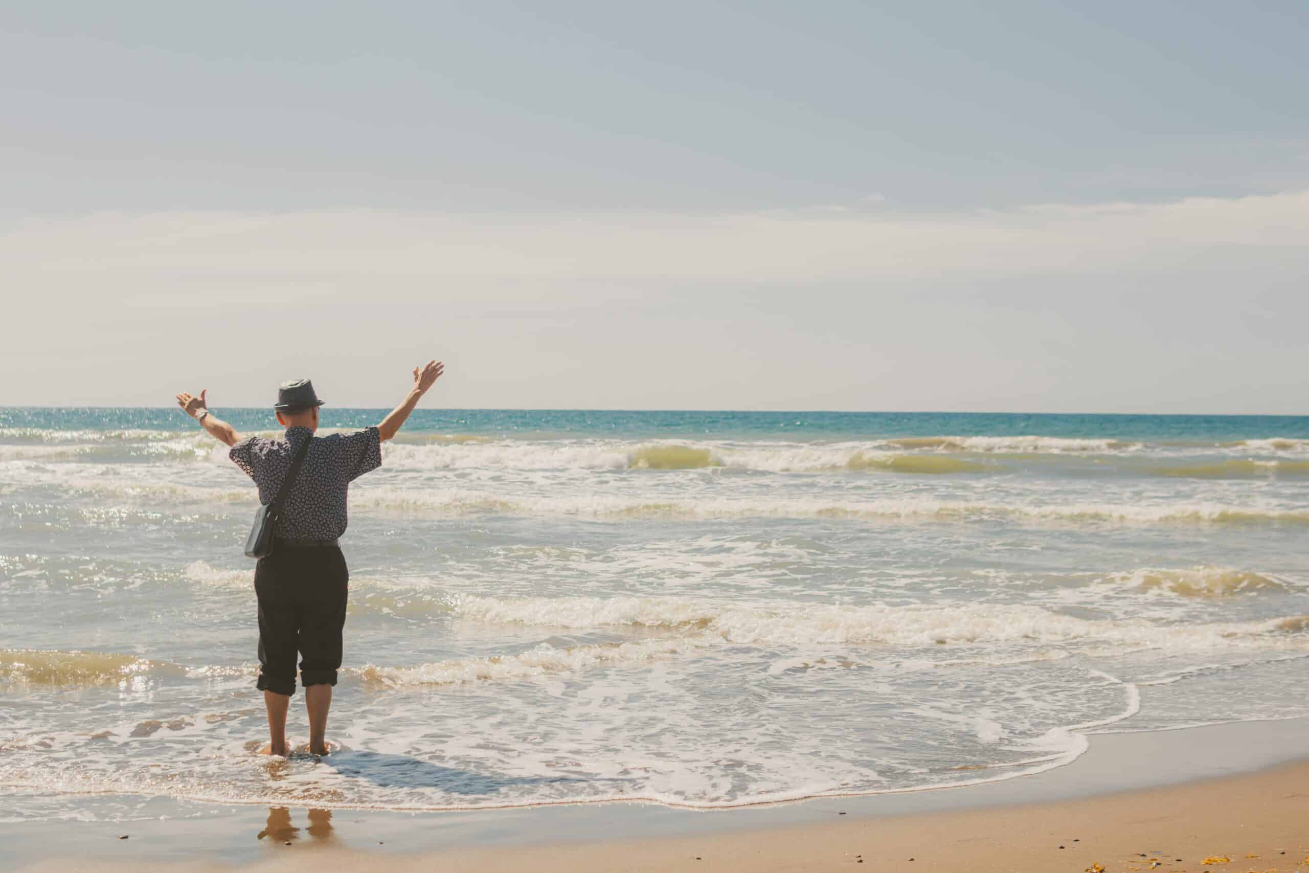Older person standing in the sea with arms up in the air, enjoying the view.