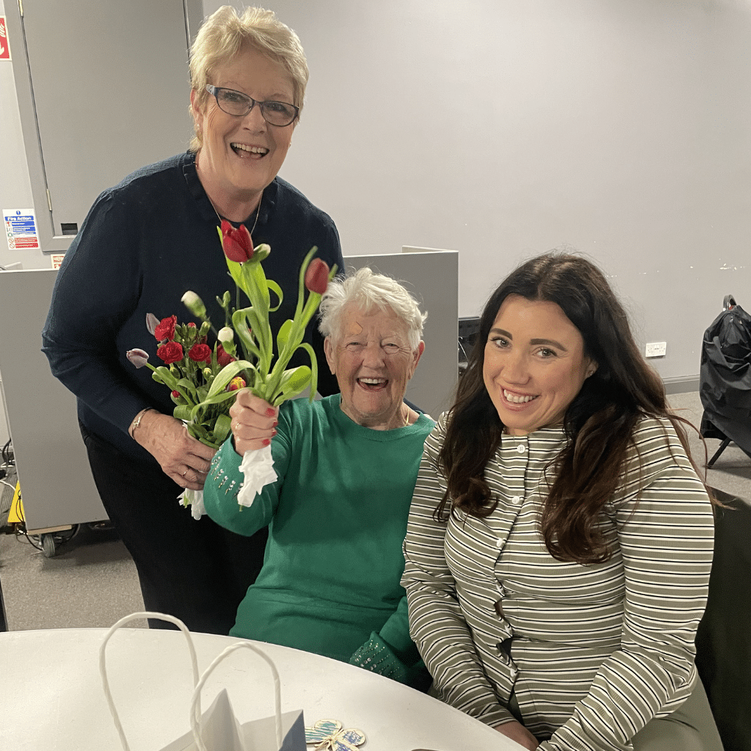service user smiling with care assistant and family member, holding a bunch of flowers