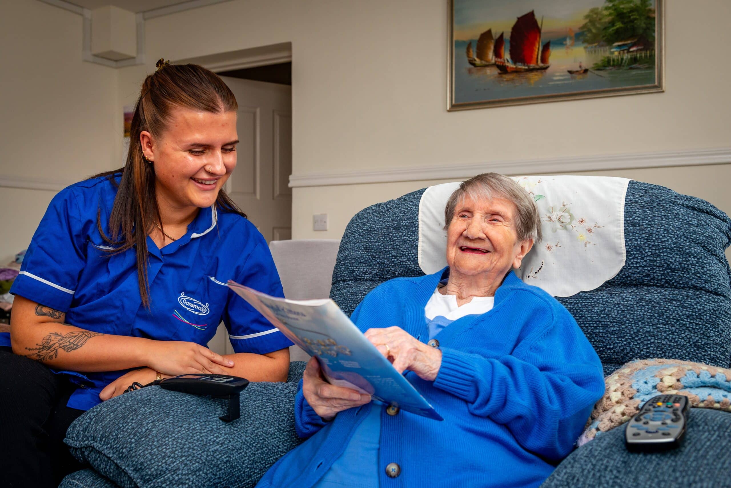 Smiling female customer sat on a chaise holding a magazine, sharing a conversation with the smiling care assistant at home