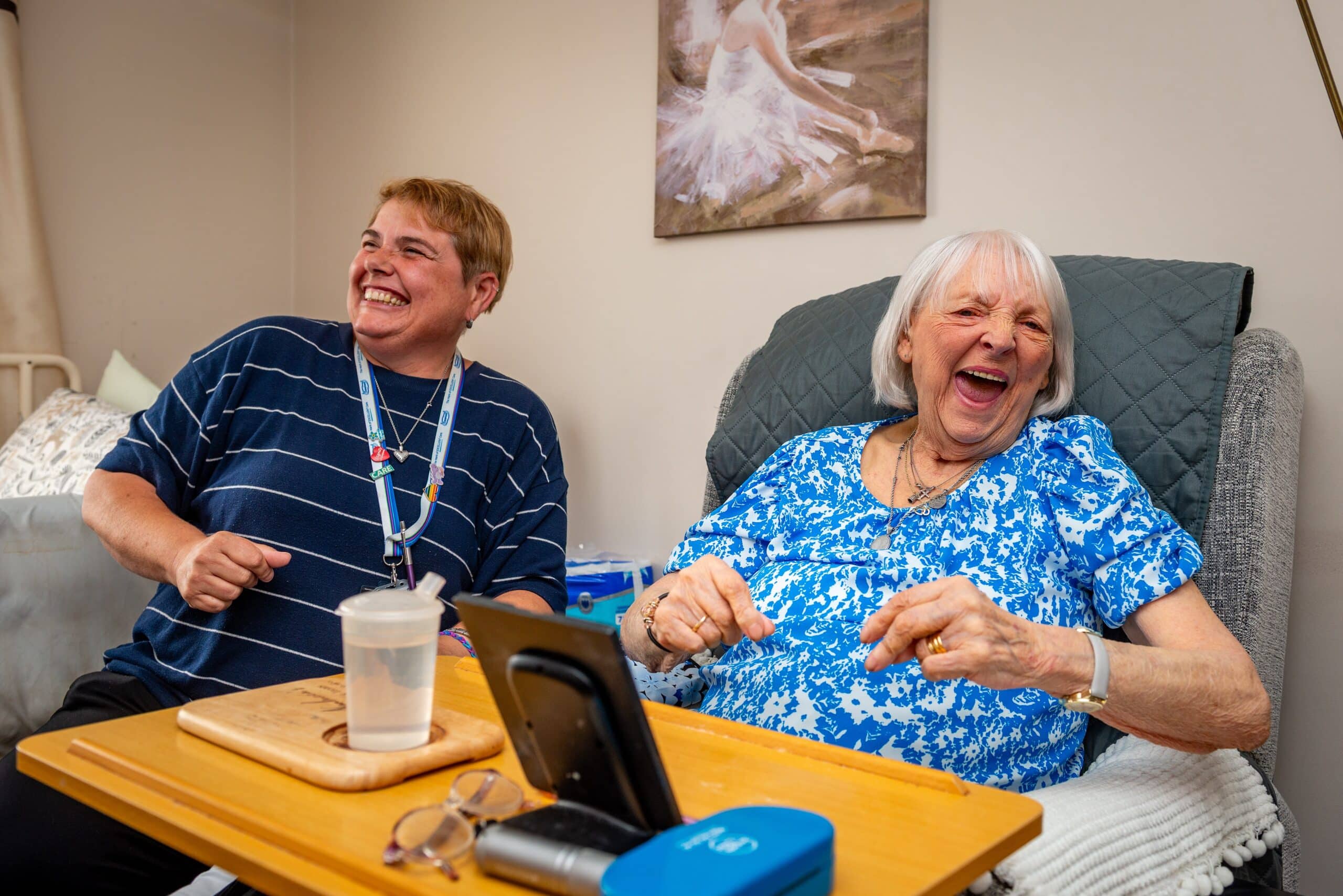 female customer receiving companionship care and laughing while sitting in her chair with a female care assistant laughing along side her